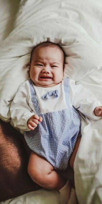 Crying baby in blue romper lying on a bed with white sheets, expressing emotion.