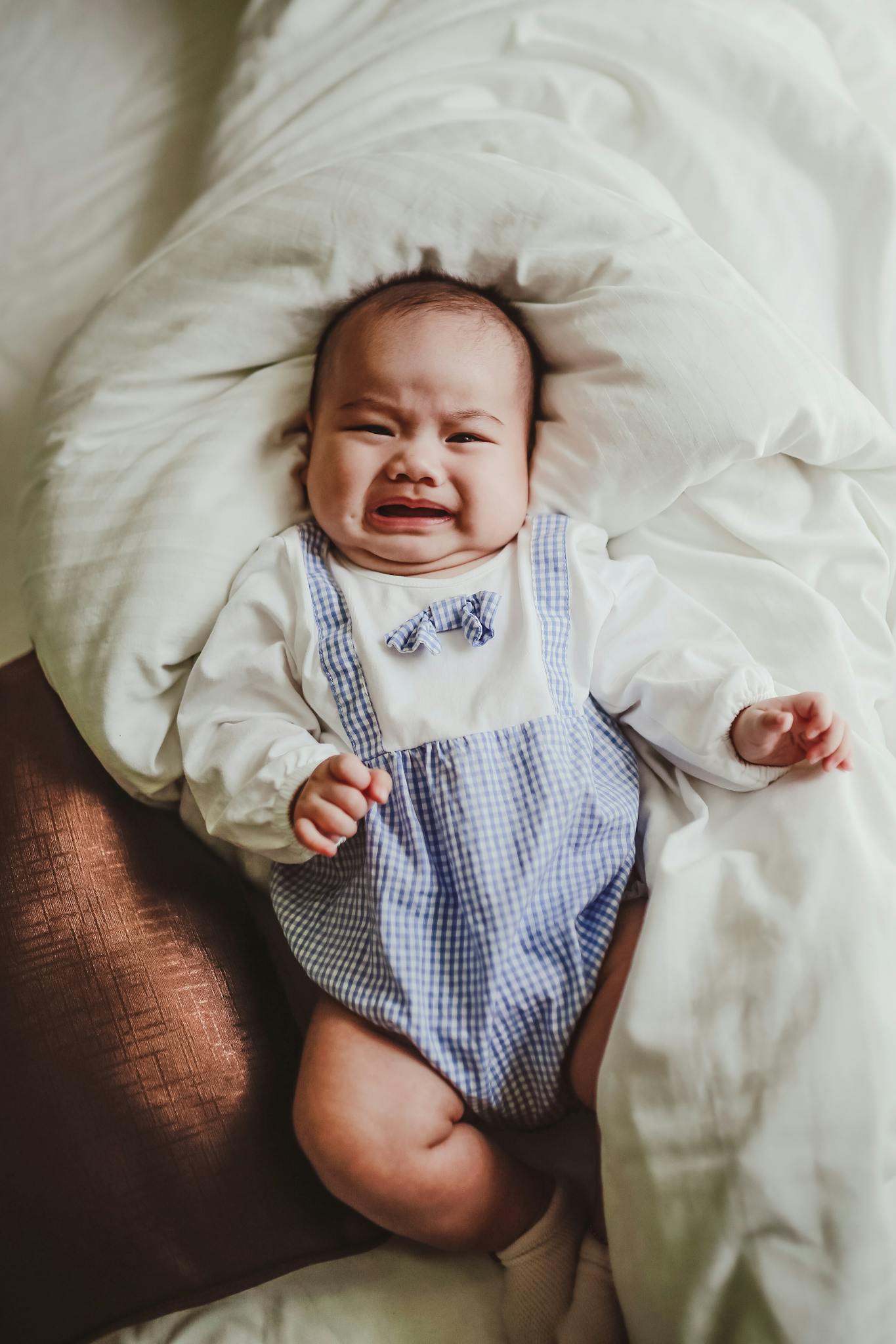 Crying baby in blue romper lying on a bed with white sheets, expressing emotion.